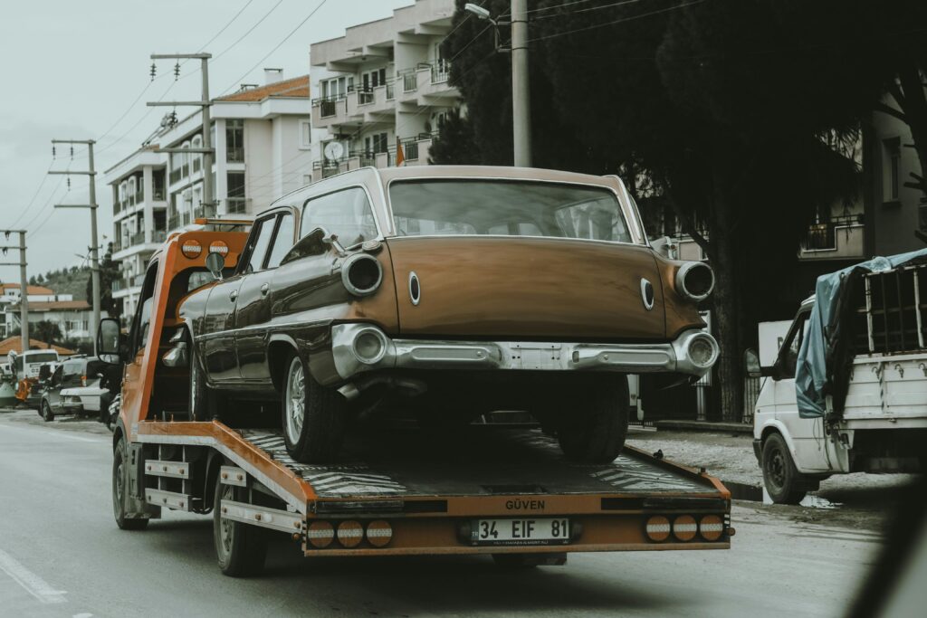 A vintage car loaded on a tow truck in a city street, surrounded by modern buildings.